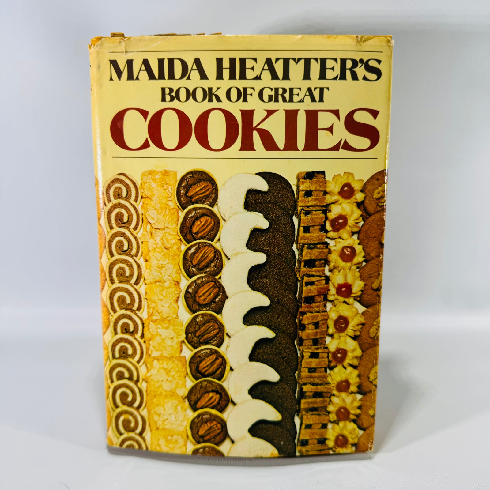 Vintage cookbook of “Maida Heatter’s Book of Great Cookies” with a cream/yellow dust jacket, bold red “COOKIES” title, and a row of assorted cookies pictured along the bottom