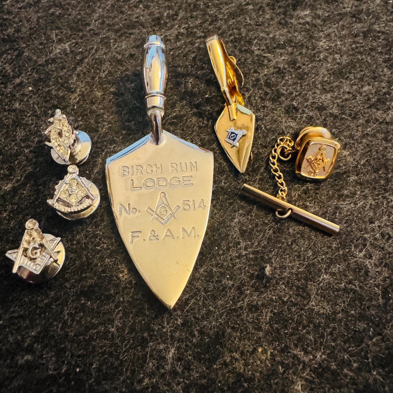 Vintage Masonic jewelry lot on black felt—Birch Run Lodge No. 514 F.&A.M. ceremonial trowel charm with handle, gold-tone Swank Masonic trowel tie clip, chain tie tack, and three Masonic lapel pins.