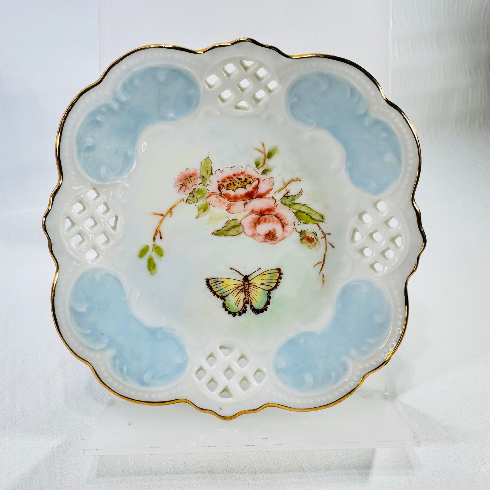 Front of a hand painted porcelain lattice dish with pink roses, a butterfly, light blue panels, and a gold scalloped rim.
