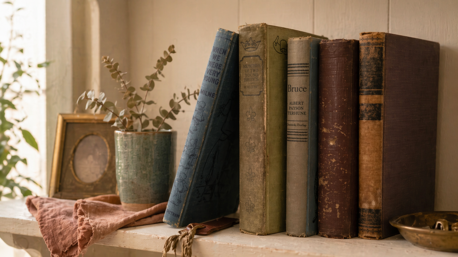 A home shelf of vintage books with a small brass picture frame, a teal ceramic cup with dried eucalyptus, and a dusty-rose linen napkin in warm late-afternoon light against an ivory wall.