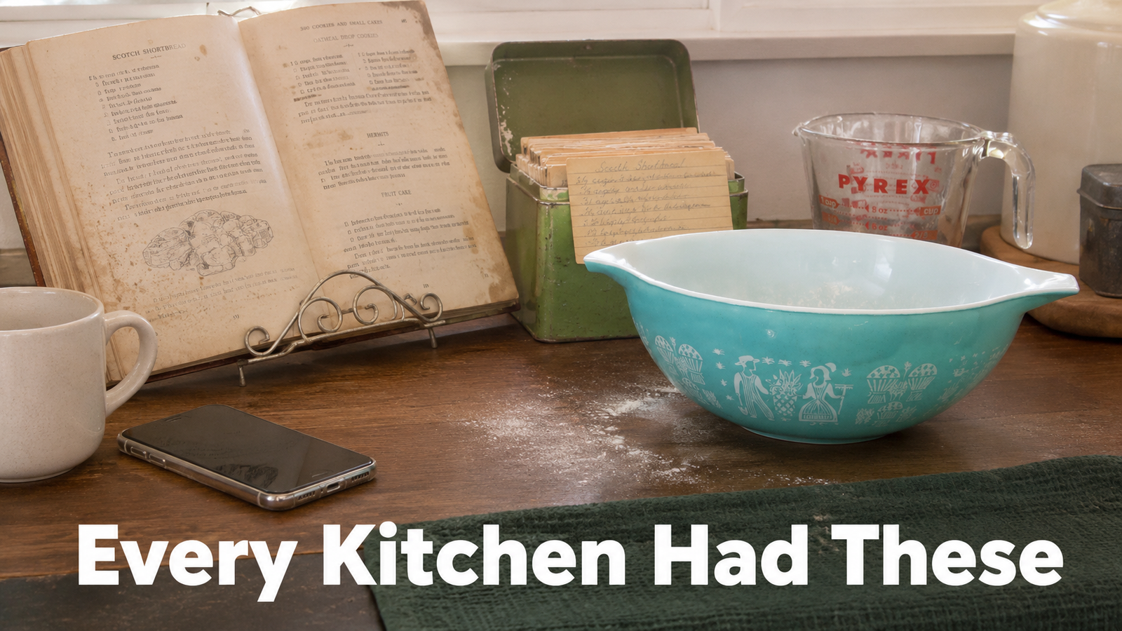 An open vintage cookbook on a metal stand beside an open 1960s green metal recipe box with handwritten cards, a patterned Pyrex Cinderella mixing bowl, and a red marked glass measuring cup on a real kitchen counter in daylight.