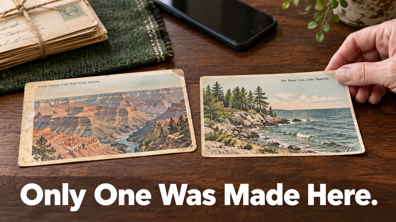  Two early 1900s American view postcards lying side by side on a dark walnut kitchen table in daylight, one showing a western canyon and one showing a Great Lakes shoreline, with a woman's hand lifting the corner of one card to turn it over.