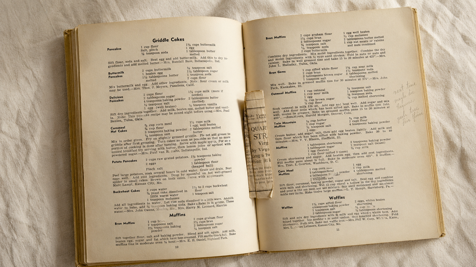 An open vintage cookbook from the 1960s showing a handwritten pencil note in the margin and a partially visible tucked-in recipe clipping, lying on warm ivory linen in soft natural light — evidence of a cookbook that was genuinely used.