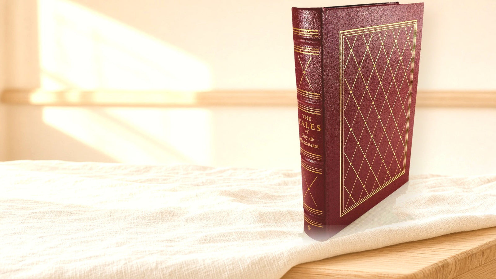 A vintage Easton Press leather-bound book standing on cream linen in soft window light, its burgundy cover stamped with a gold lattice pattern.