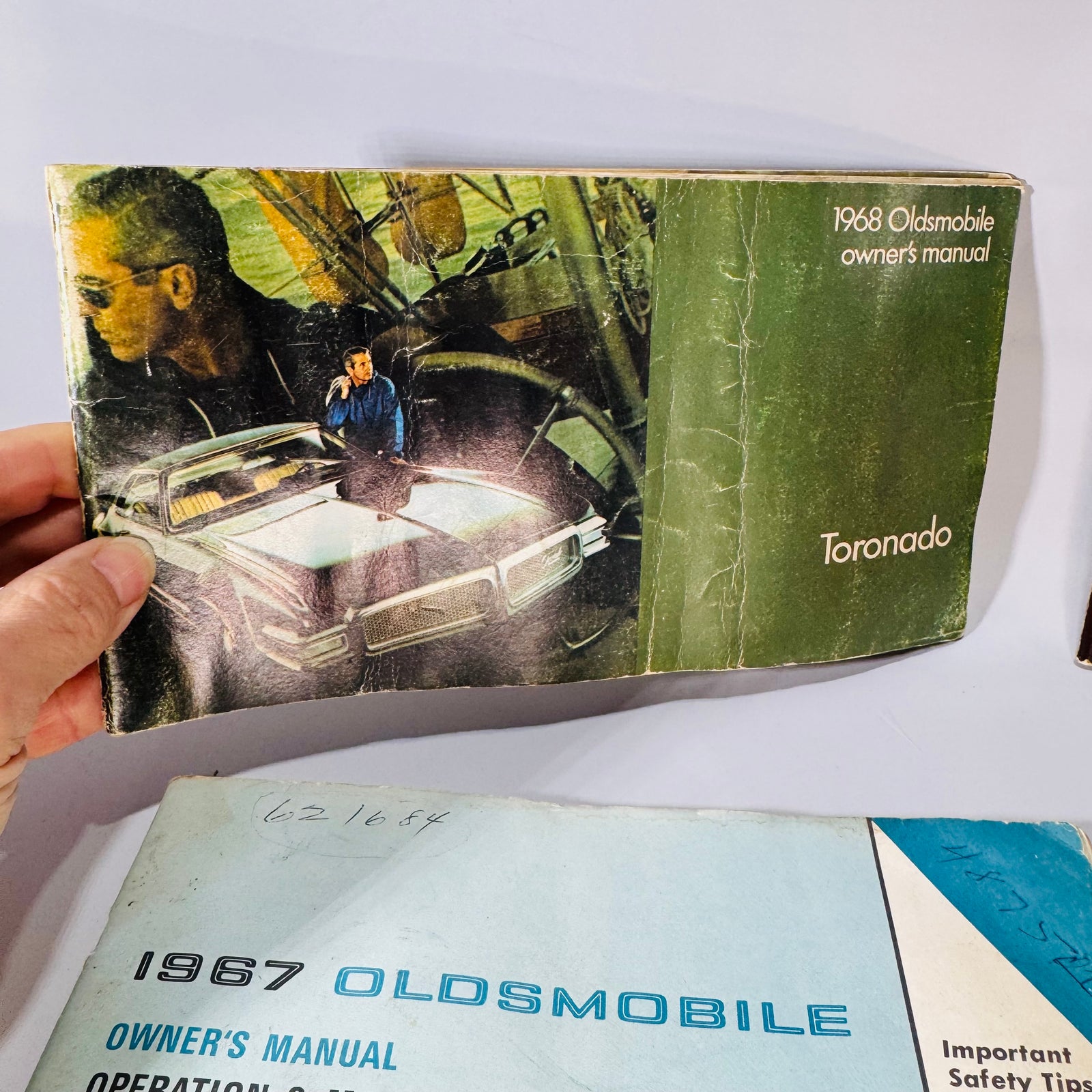 Vintage Oldsmobile paper bundle laid out on a wood surface—1968 Toronado Owner’s Manual (green), 1967 Toronado Owner’s Manual (red), 1967 Operation & Maintenance Instructions booklet (blue, smaller), and 1967 Oldsmobile SPECS booklet—showing visible wear, water staining, and handwritten notes.