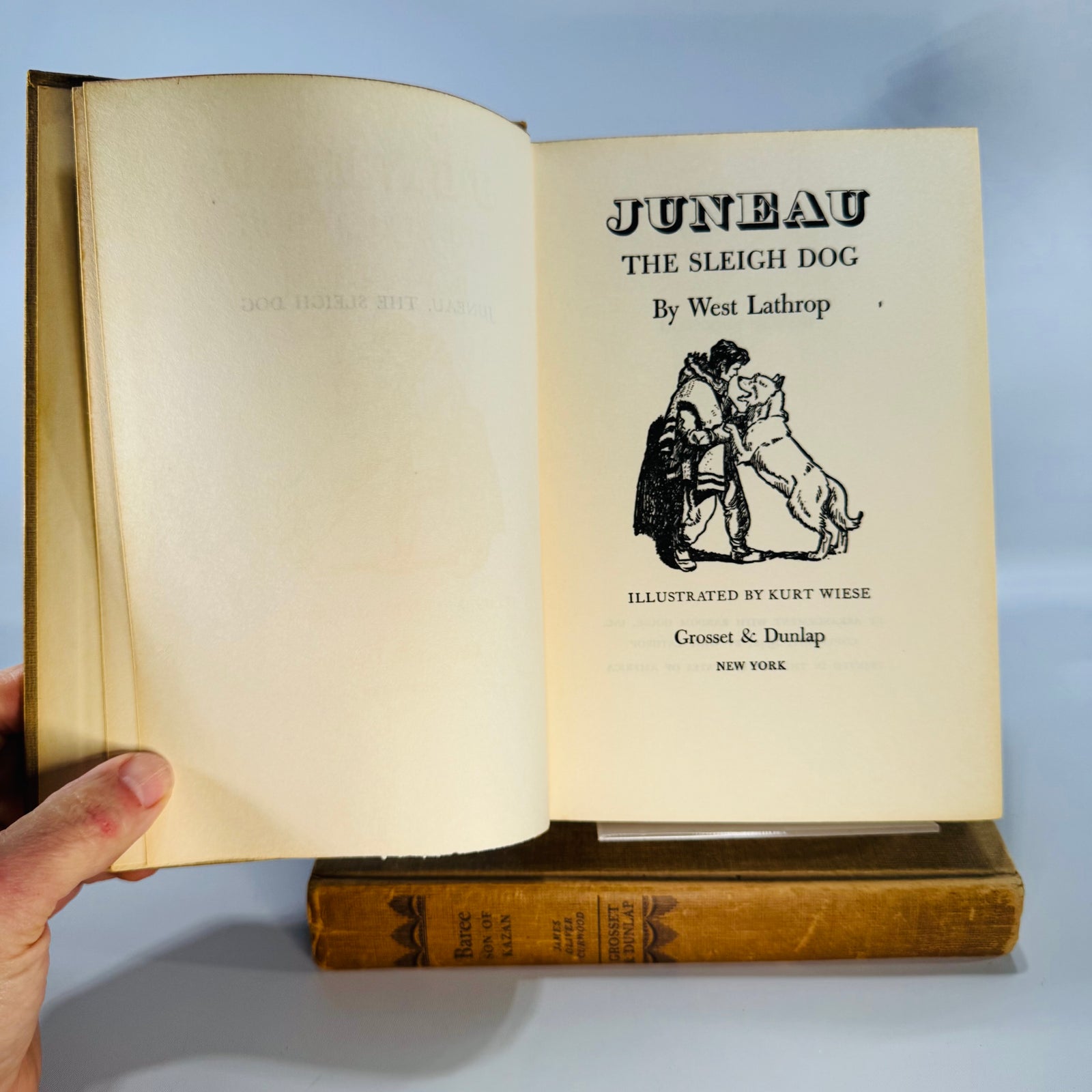 Two vintage tan cloth dog-story hardcovers—Baree: Son of Kazan and Juneau: The Sleigh Dog—standing side by side with worn spines on a white backdrop.