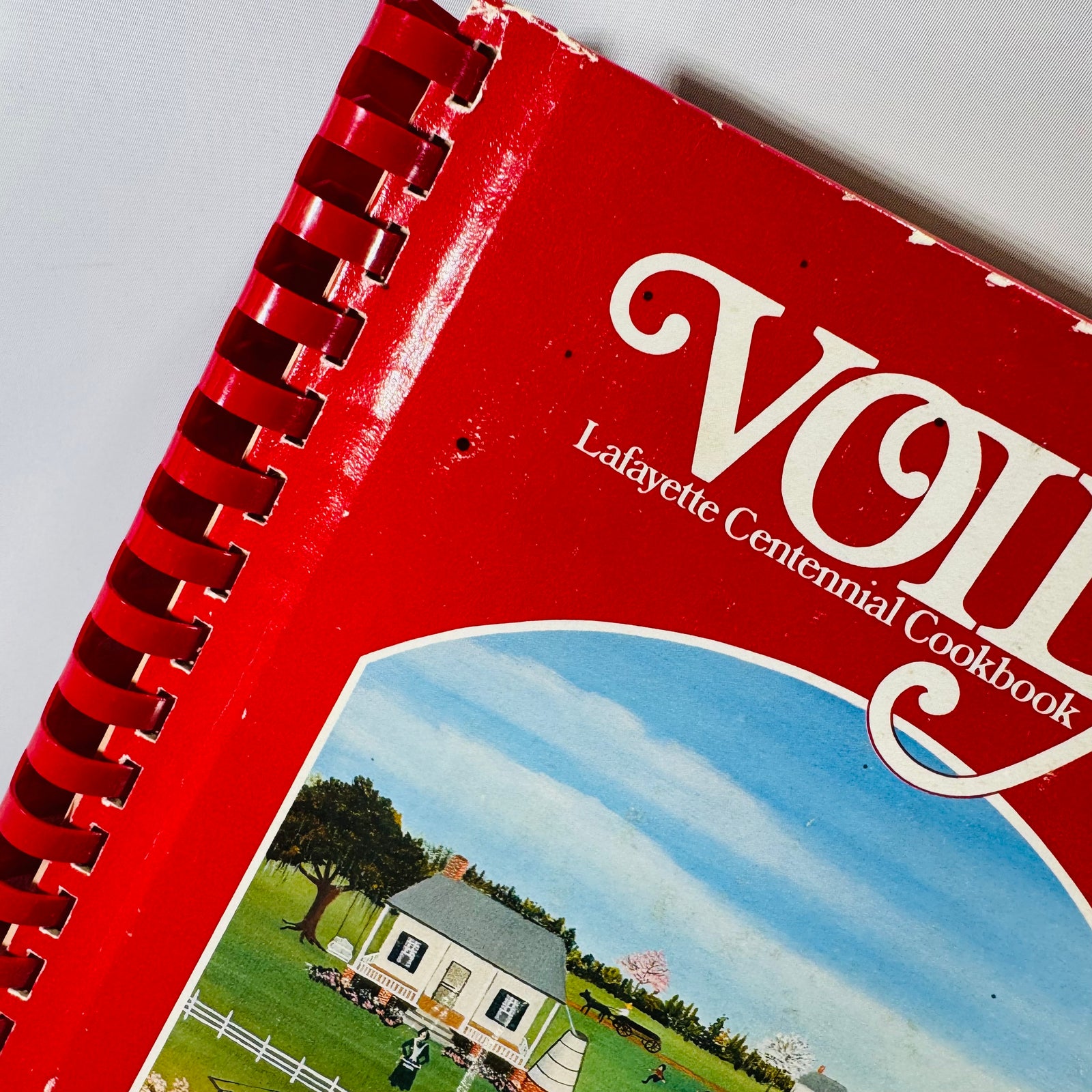 Vintage cookbook bundle on a white backdrop: red spiral-bound “VOILA! Lafayette Centennial Cookbook 1884–1984” beside red comb-bound “Culinary Cues,” with “Cora’s Country Cookbook” featuring a woman holding a pie placed in front.