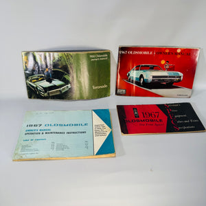 Vintage Oldsmobile paper bundle laid out on a wood surface—1968 Toronado Owner’s Manual (green), 1967 Toronado Owner’s Manual (red), 1967 Operation & Maintenance Instructions booklet (blue, smaller), and 1967 Oldsmobile SPECS booklet—showing visible wear, water staining, and handwritten notes.