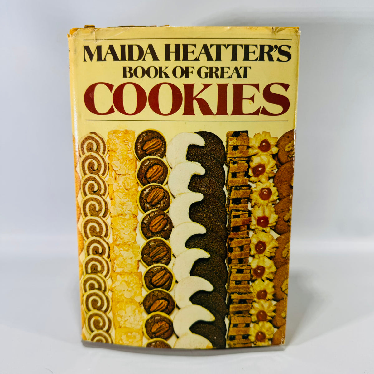 Vintage cookbook of “Maida Heatter’s Book of Great Cookies” with a cream/yellow dust jacket, bold red “COOKIES” title, and a row of assorted cookies pictured along the bottom