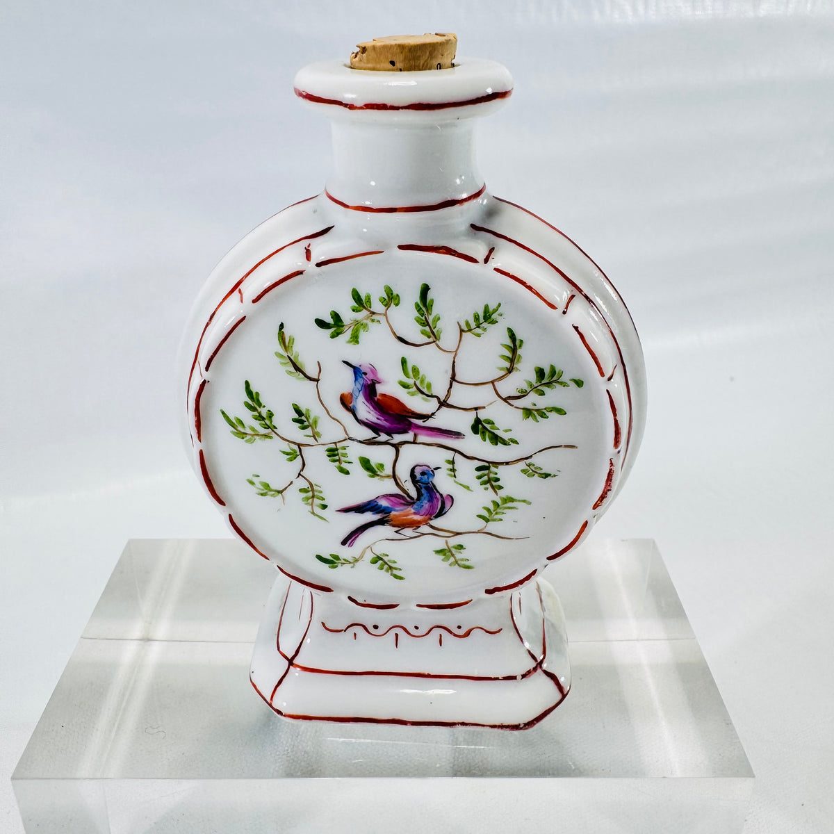 Front of a hand painted porcelain moon flask bottle with two birds on leafy branches, red trim, and cork stopper.