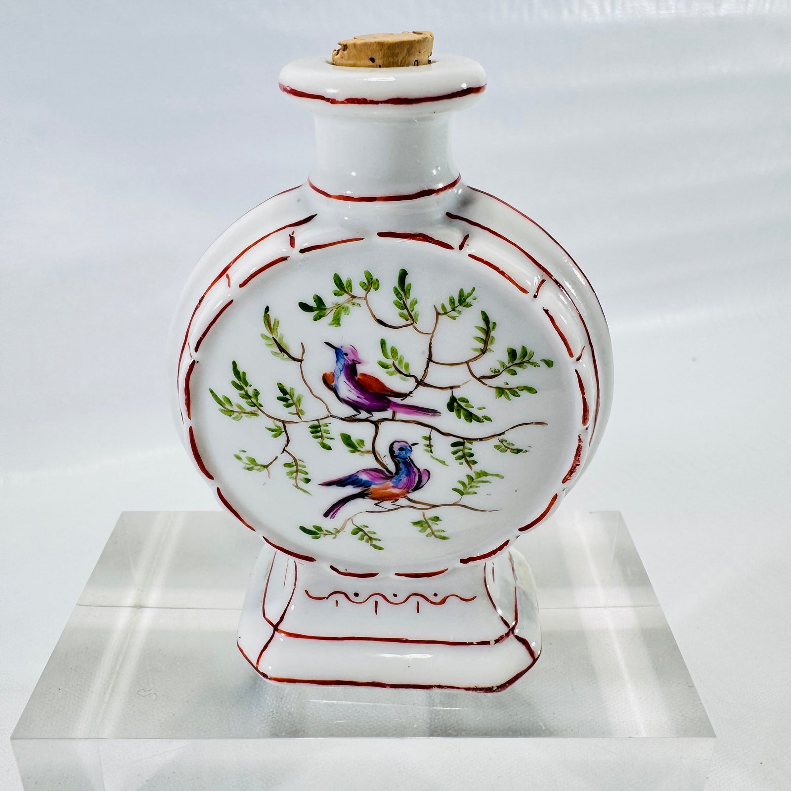 Front of a hand painted porcelain moon flask bottle with two birds on leafy branches, red trim, and cork stopper.