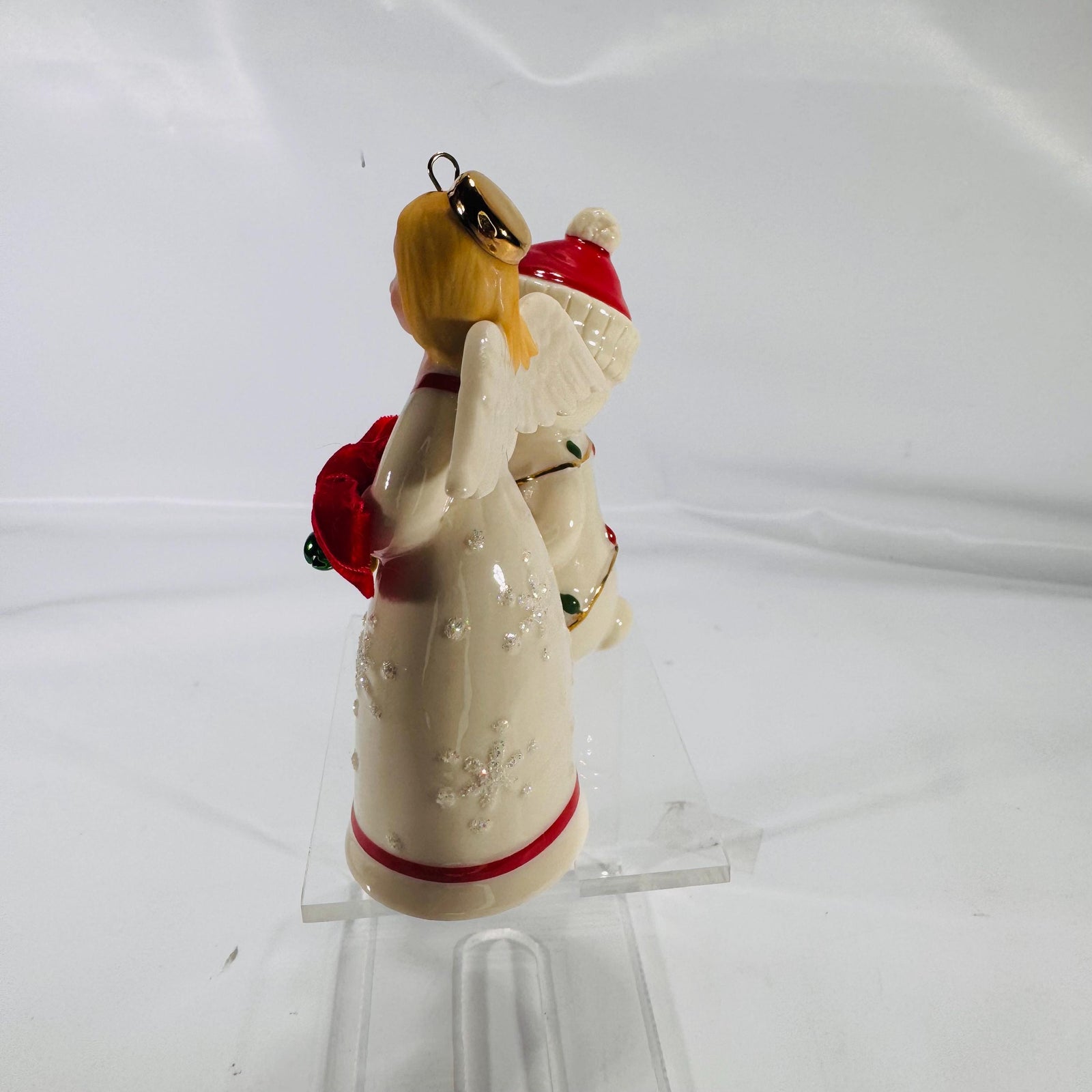 Front view of two Lenox® Christmas figurines on a clear acrylic stand—a 4″ porcelain teddy bear wrapped in gold-trimmed string lights wearing a red Santa hat next to a 4″ porcelain angel holding jingling bells with a red velvet bow, both with gold accents.