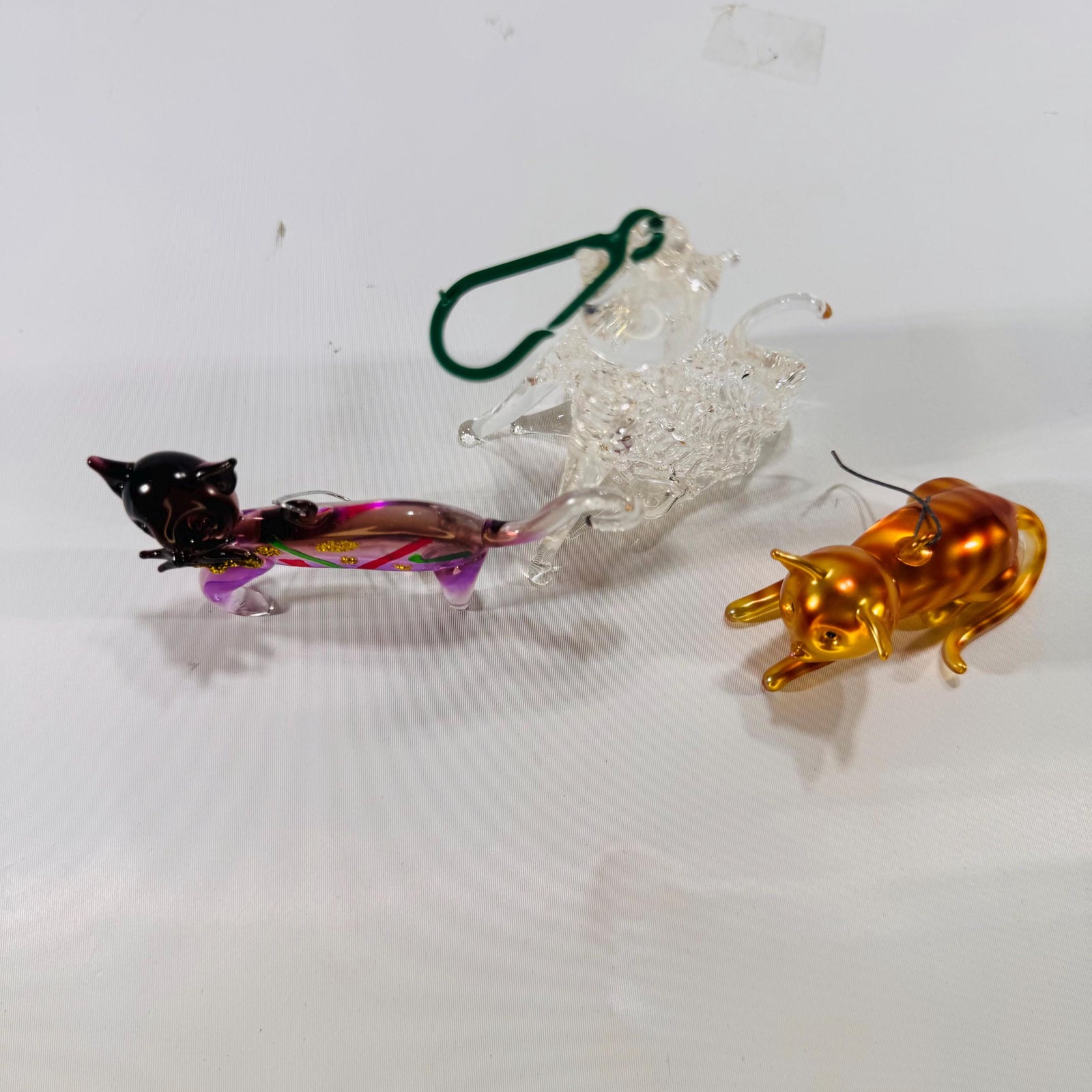 Three handblown glass cat ornaments—left: purple cat with red and green accents and gold glitter; center: clear crackle-glass cat sitting upright; right: metallic gold-finish cat in a crouching pose