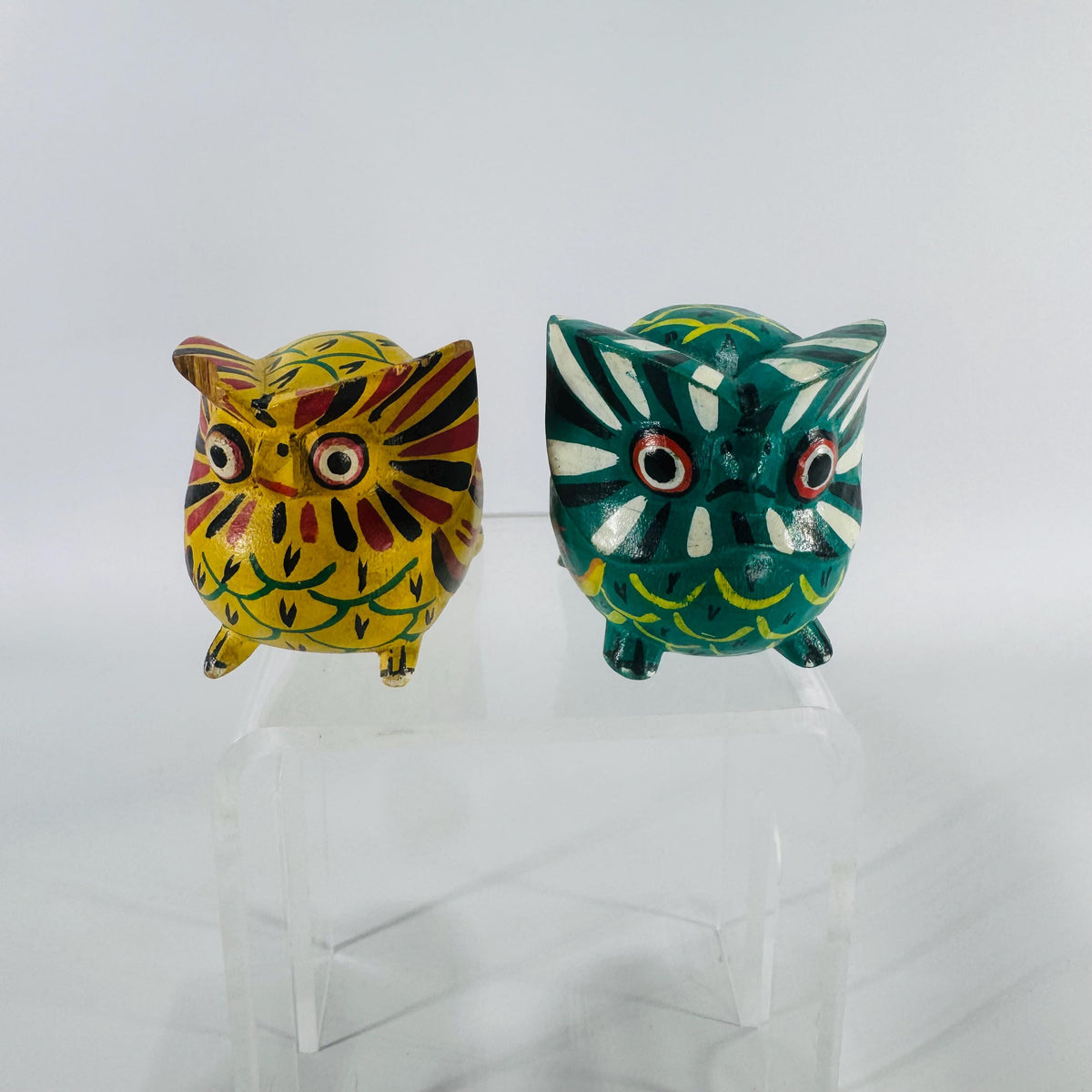 Two small hand-painted wooden owl figurines—one teal and one yellow—with bold folk-art patterns, shown front facing on a white background.
