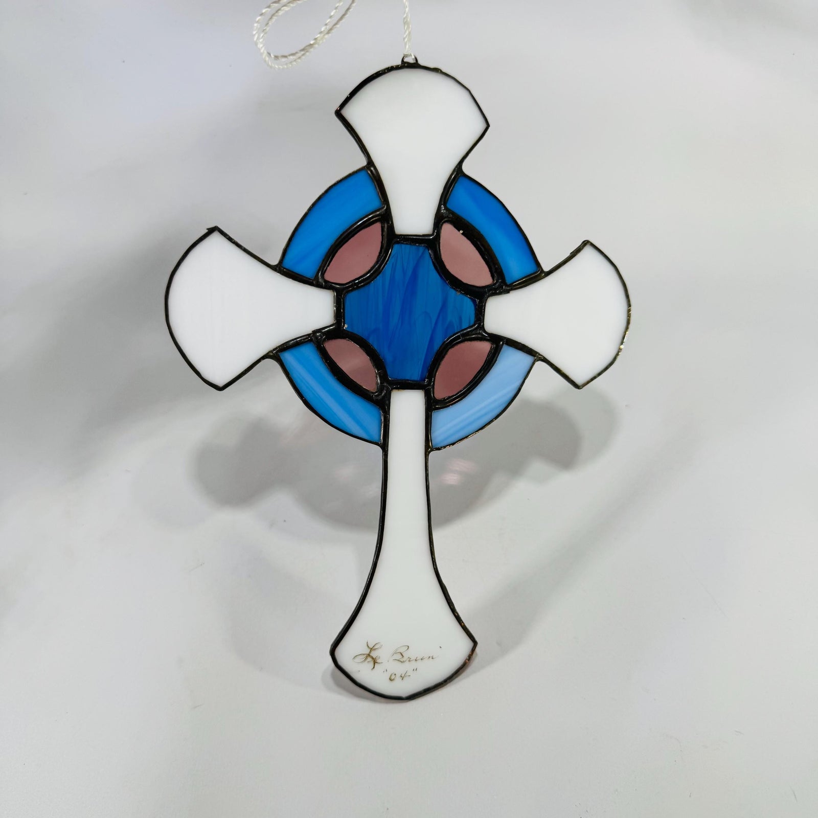 Front view of stained-glass Celtic cross with blue ring and white arms, hanging by white cord on a white background.