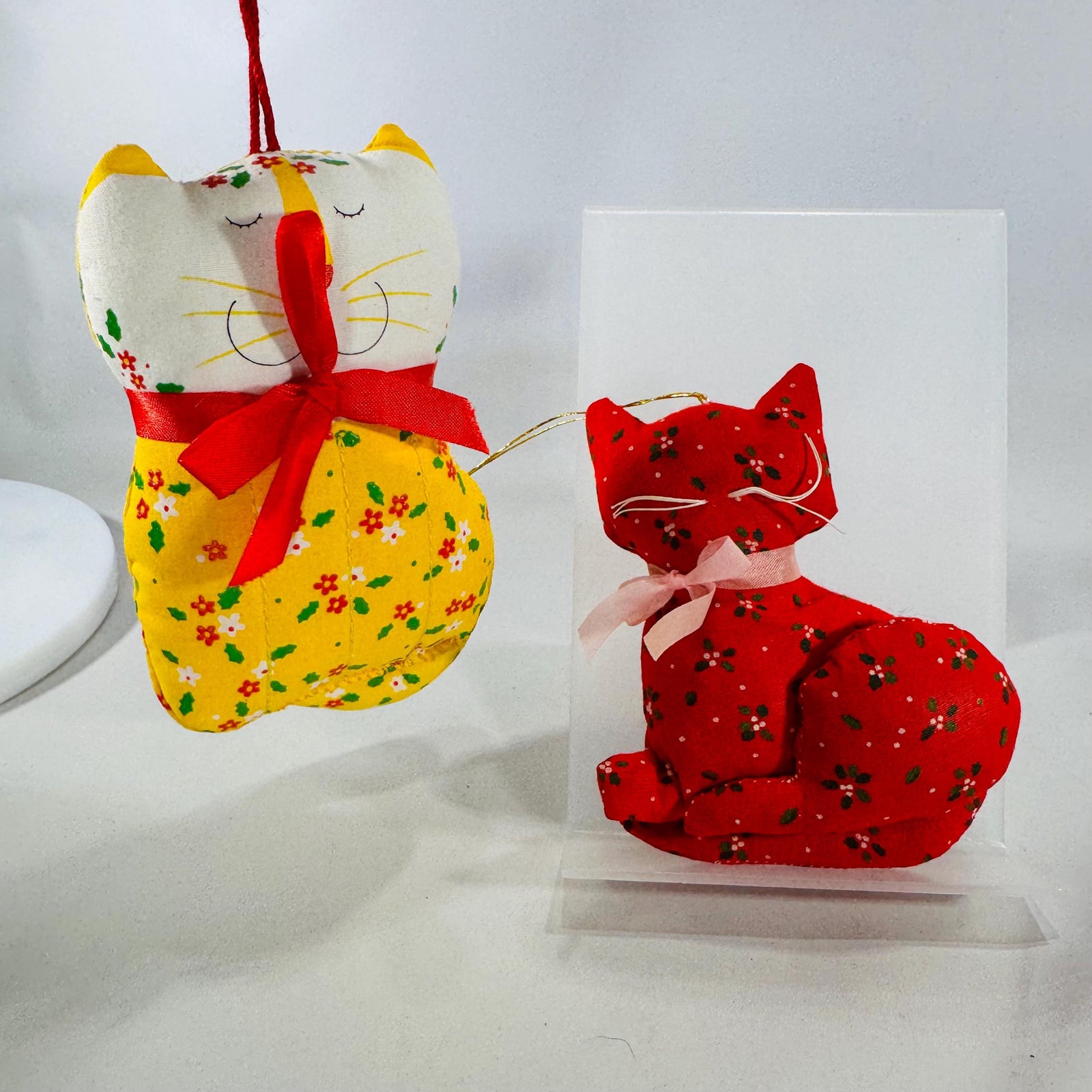 Two 1970s–80s handmade fabric cat ornaments, one yellow and one red, with floral prints and ribbon bows.