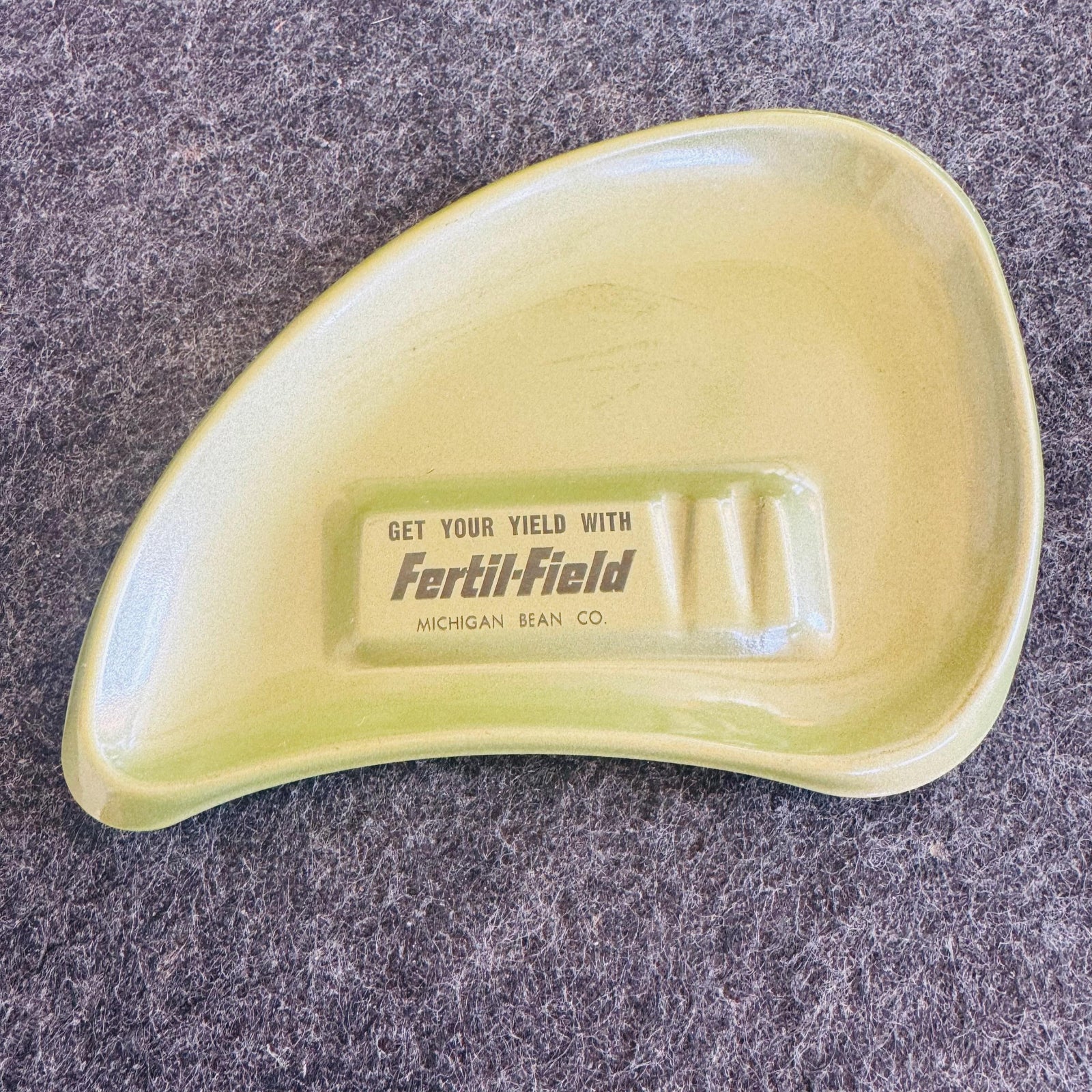Vintage avocado green boomerang ashtray with black “Get your yield with Fertil-Field” Michigan Bean Co advertising printed in the center.