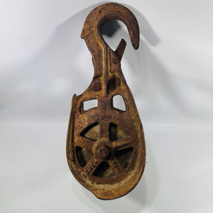 Vintage cast iron single-wheel barn pulley with large fixed hook and safety latch, viewed from the front, showing open spoked iron wheel frame, deep rust and ochre patina throughout, a