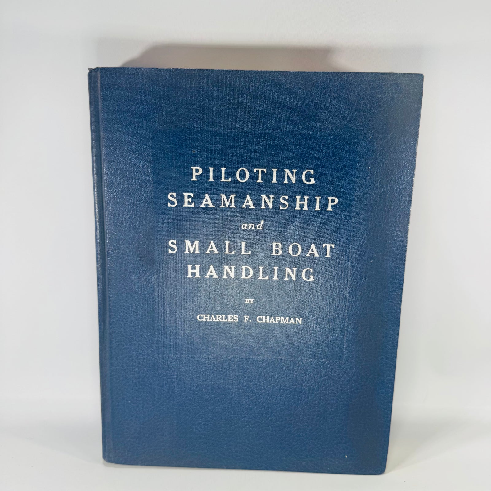 Navy blue hardcover vintage boating manual titled “Piloting, Seamanship and Small Boat Handling” by Charles F. Chapman, with interior photos of nautical charts and seamanship spreads.