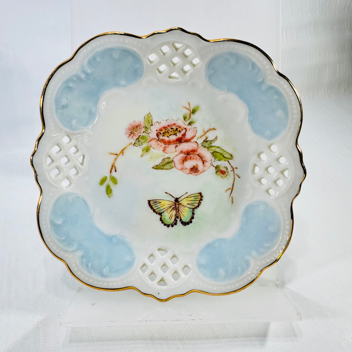Front of a hand painted porcelain lattice dish with pink roses, a butterfly, light blue panels, and a gold scalloped rim.