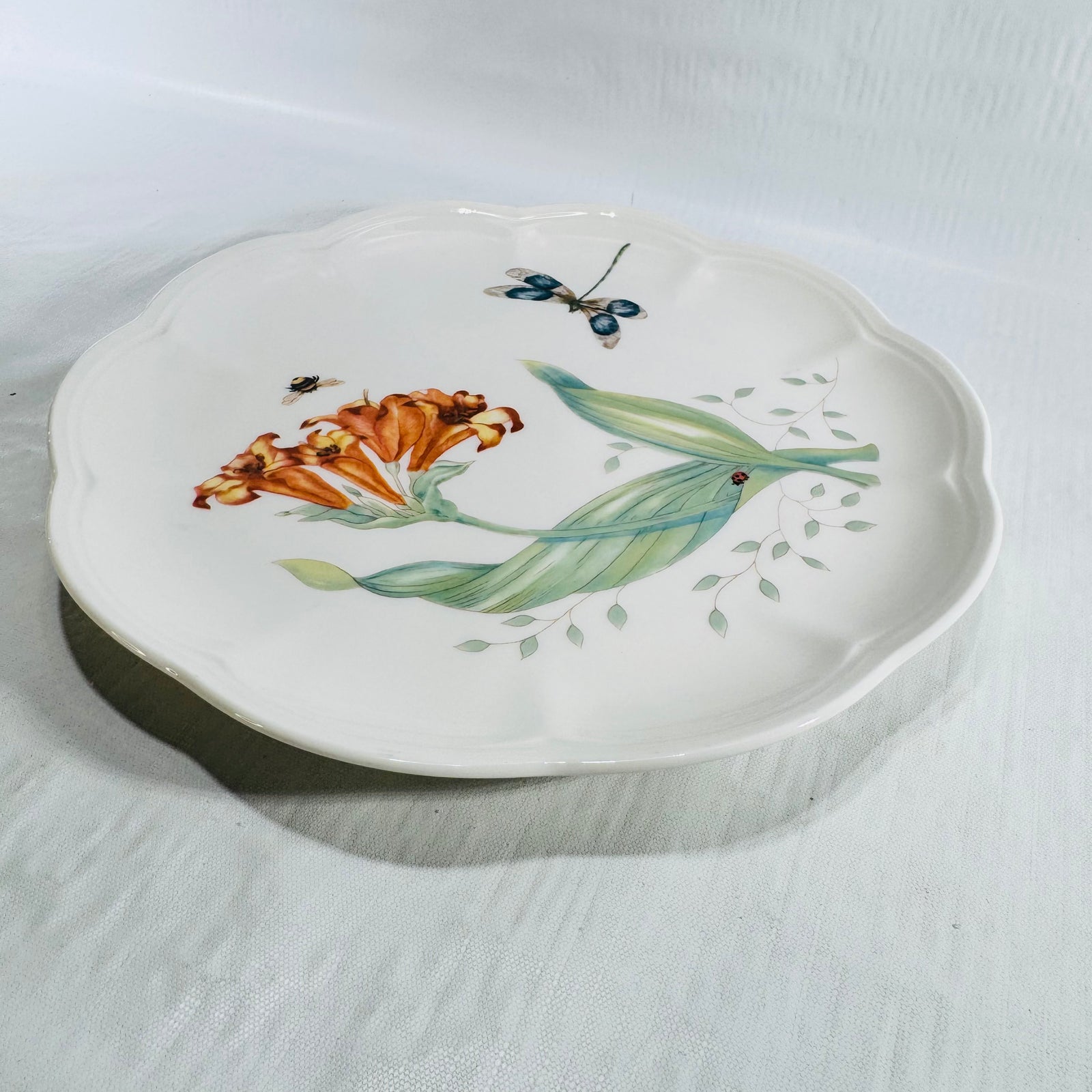 Angled view of Lenox Butterfly Meadow Dragonfly 9 inch plate on white fabric with orange lilies