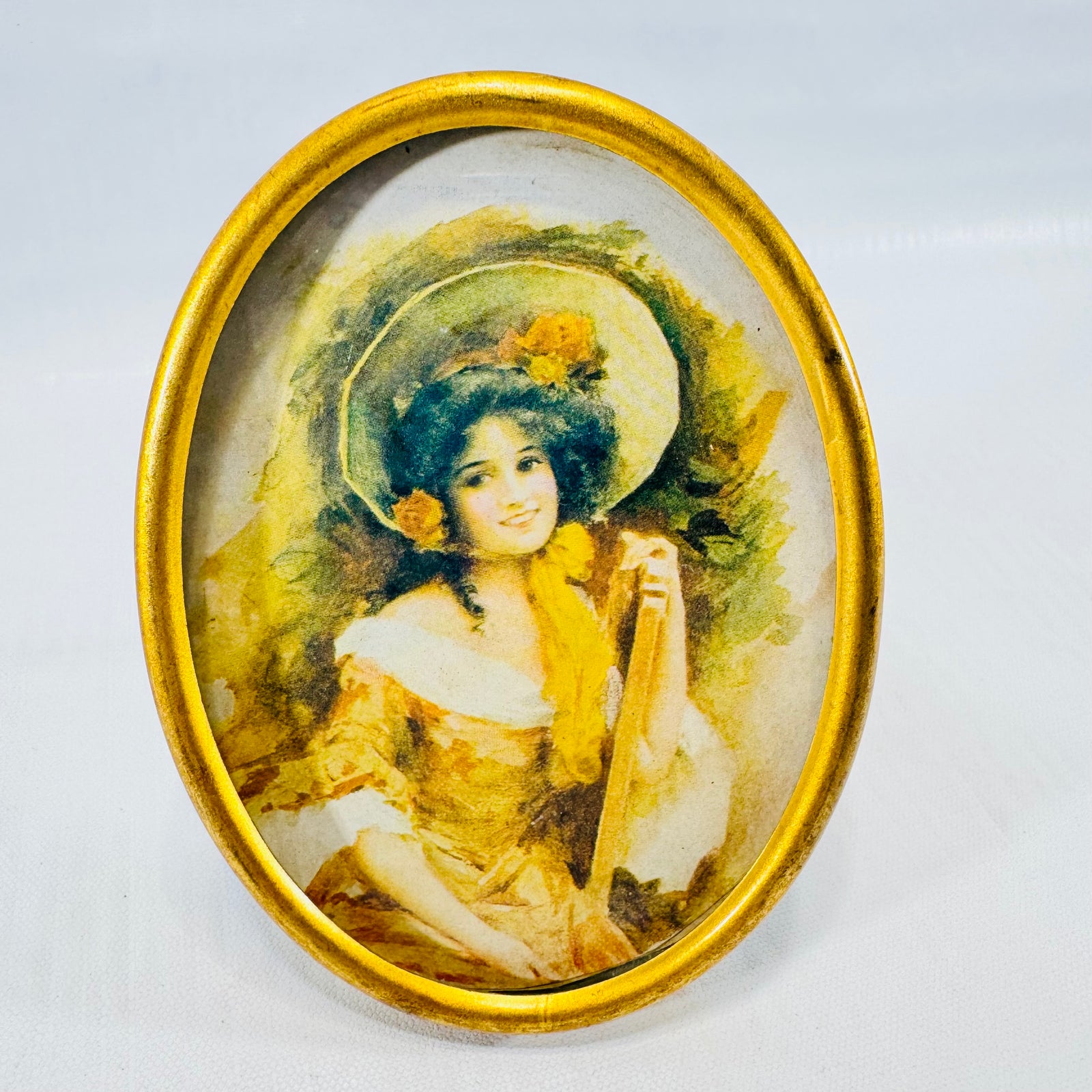 Front of a 4 inch oval gold tone easel frame holding a print of a smiling woman in a large flower bonnet.