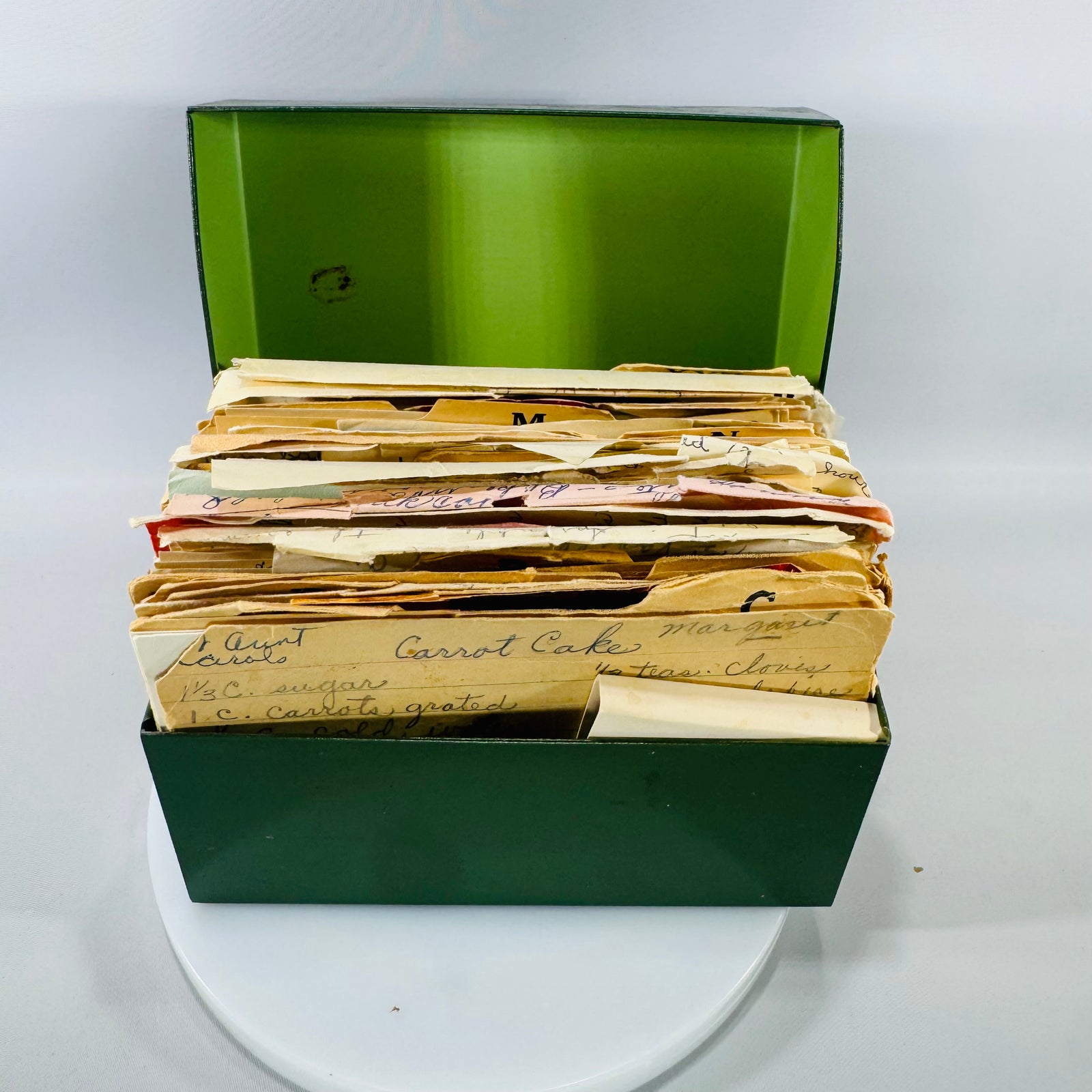 Deep green vintage metal recipe box opened to reveal a thick stack of handwritten recipe cards and newspaper/magazine clippings, including “Aunt Carol’s Carrot Cake.”
