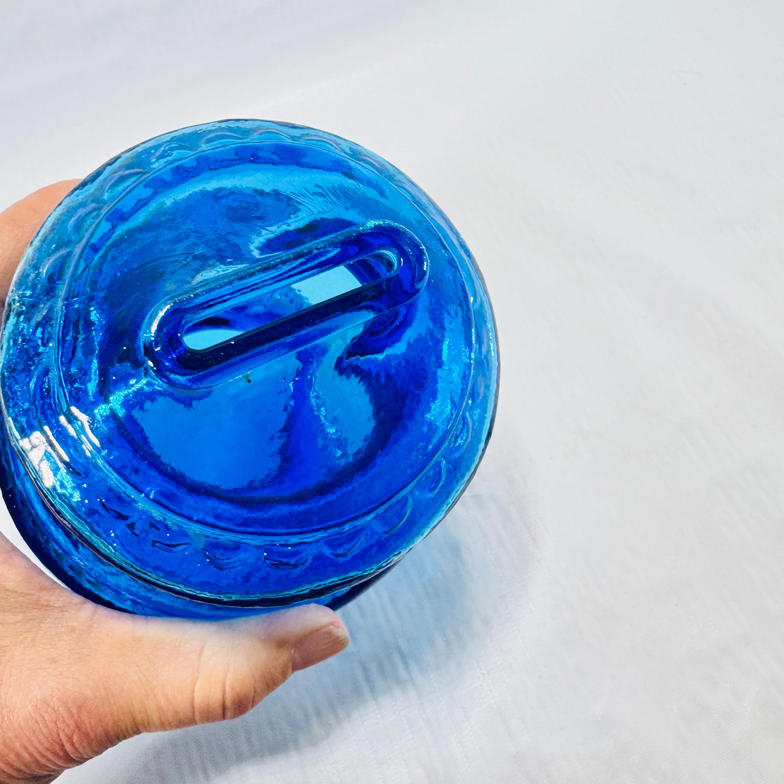 Coin slot on top of the blue glass bank held in hand, with a tiny speck visible inside.