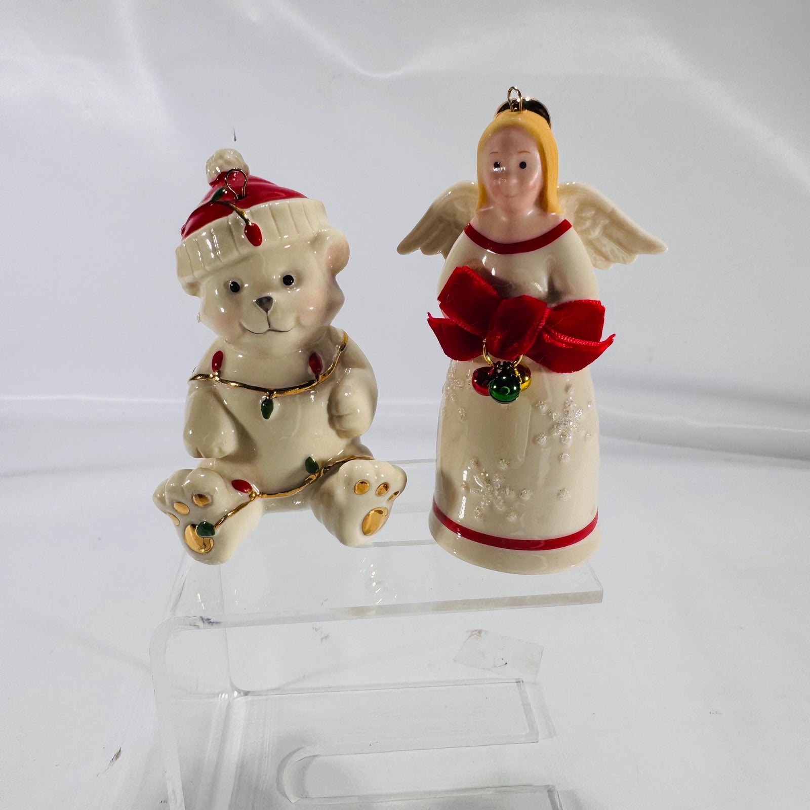 Front view of two Lenox® Christmas figurines on a clear acrylic stand—a 4″ porcelain teddy bear wrapped in gold-trimmed string lights wearing a red Santa hat next to a 4″ porcelain angel holding jingling bells with a red velvet bow, both with gold accents.